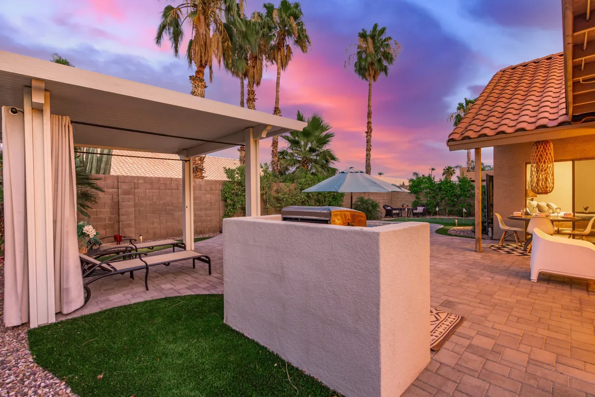 The backyard showcases a comfortable outdoor seating area, barbecue grill, lush palm trees, and a colorful sunset. The setting is ideal for relaxation or casual gatherings after a busy day.