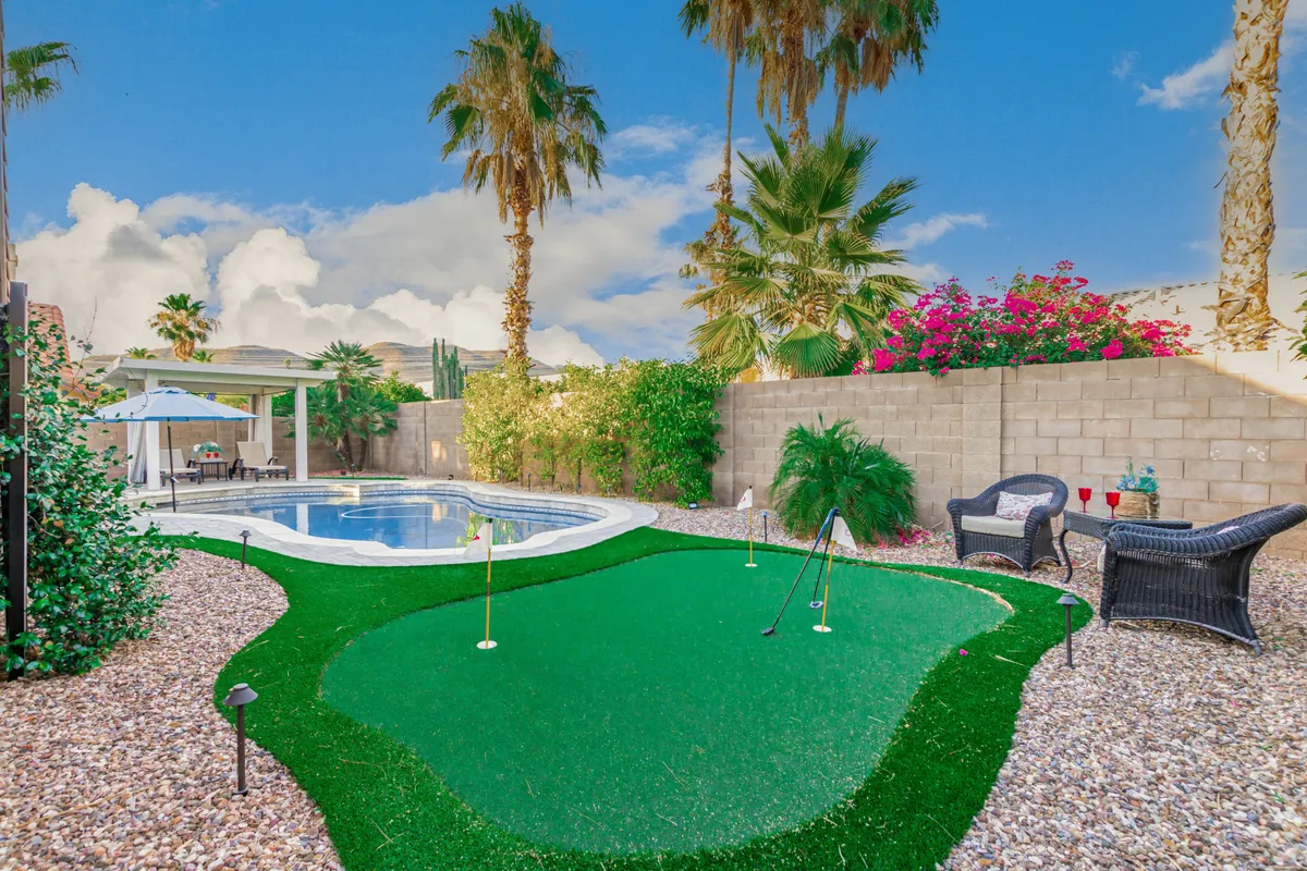 This exterior image prominently features an artificial putting green just steps from a swimming pool, surrounded by palm trees, lounge chairs, and flowering shrubs. The scene is lively yet relaxing, suggesting both golf practice and poolside leisure in a lush backyard environment.