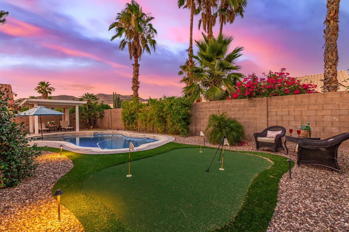 A sweeping view of the backyard highlights the sparkling swimming pool, a lush golf putting green, tall palm trees, and inviting lounge chairs surrounded by flowering plants. The scene exudes luxury and tranquility, showcasing the opportunities for swimming, golf practice, and poolside relaxation.