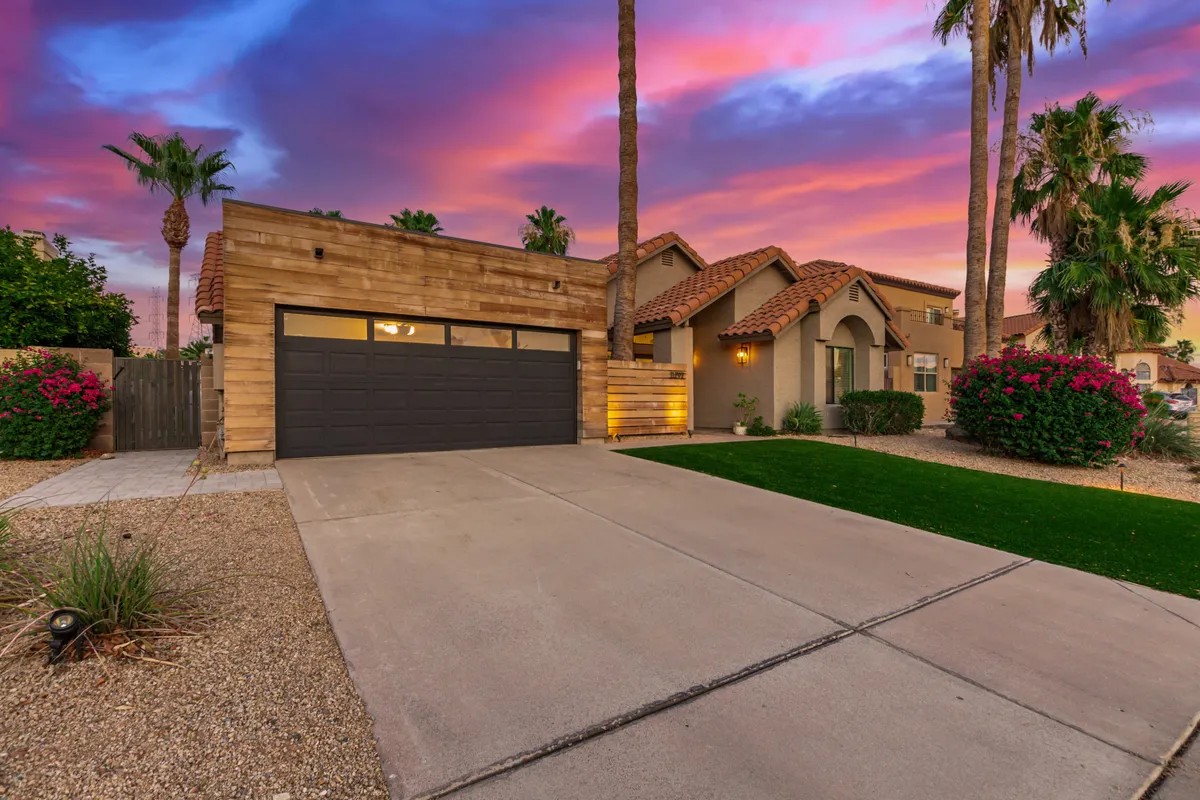 This exterior shot features a modern garage door framed by elegant palm trees and a vibrant sunset sky. Decorative landscaping softens the entrance, and the scene conveys a sense of arrival and tranquility.
