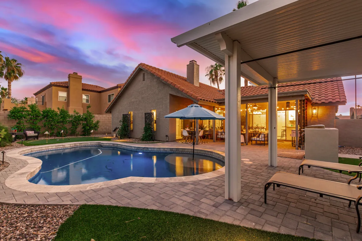 A lively exterior scene featuring a sparkling swimming pool surrounded by a patio area with stylish outdoor furniture, a dining space, and lush landscaping. The inviting water and ample seating set the stage for poolside fun and family gatherings.