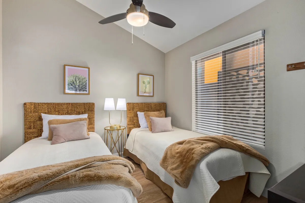 A comfortable guest bedroom with two beds featuring woven headboards, decorative pillows, and warm lamp lighting. Framed artwork and sunlight streaming in create an inviting, restful environment, well-suited for multiple guests.