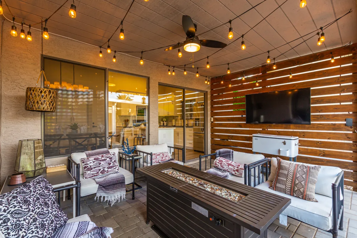 An inviting outdoor area with a fire pit, cozy furniture, hanging lights, and a mounted TV creates a space for relaxation and socializing into the evening.