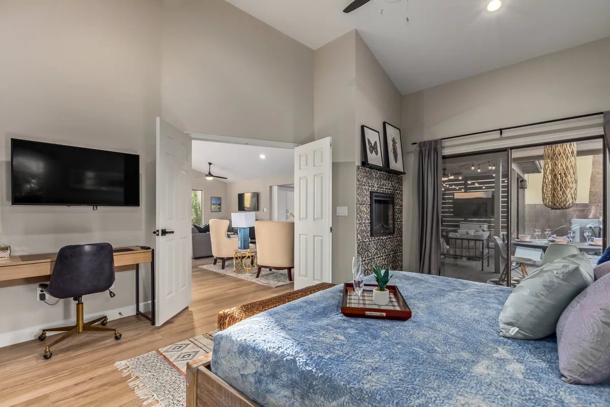 A comfortable bedroom with blue bedding, a fireplace, a work desk, and a view into the living area. The space is bright and inviting, offering areas for rest, work, and relaxation.