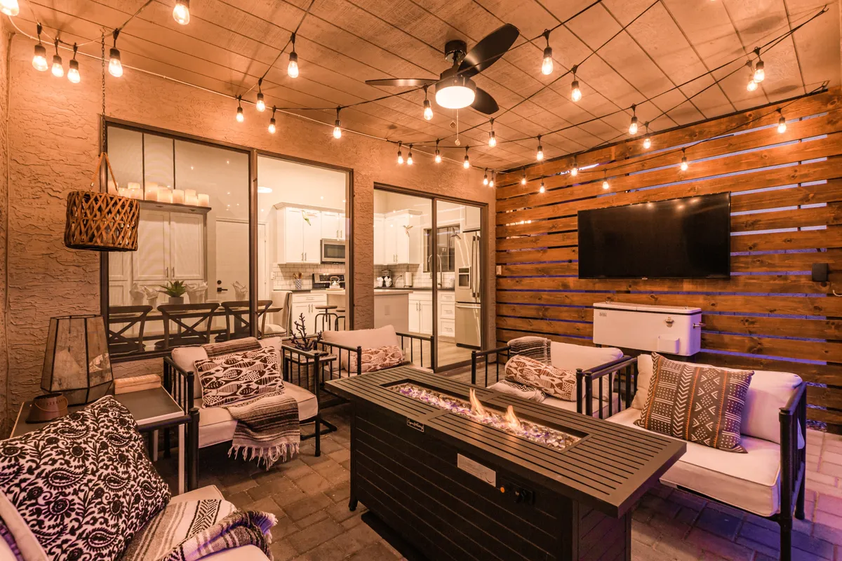 An intimate outdoor living area featuring a modern fire pit, hanging string lights, a wooden accent wall, and plush seating arranged for conversation. The outdoor TV is visible, creating a perfect space for sunset viewing or movie nights.