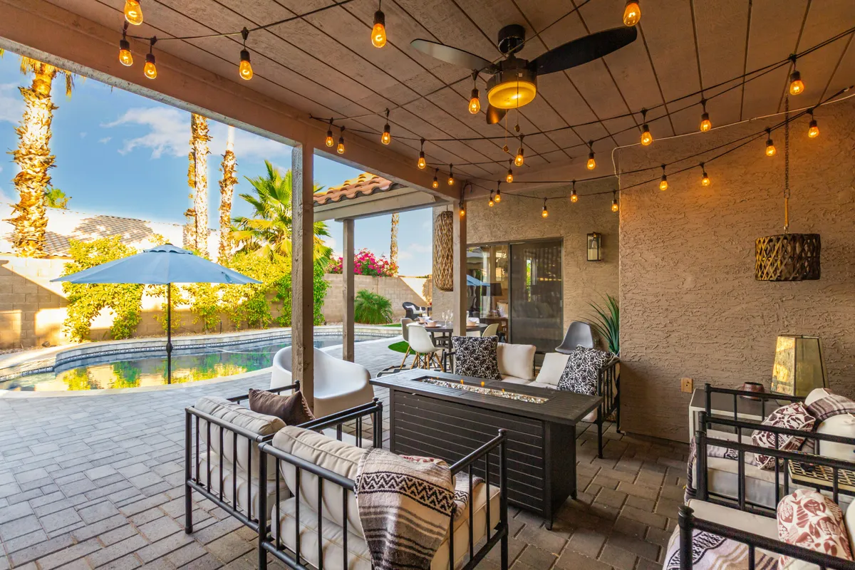 The image shows a cheerful exterior setting with a pool, outdoor seating, string lights, umbrella, and potted plants. The area is ideal for evening relaxation, pool parties, or social gatherings, with a cozy, inviting vibe as dusk falls.