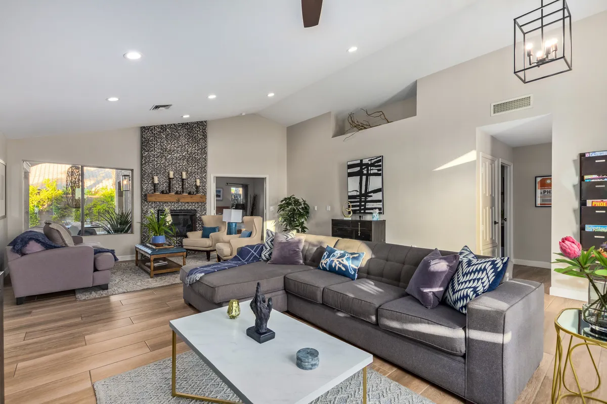 A spacious living room with a large sectional sofa arranged around a coffee table, a decorative tiled fireplace, and stylish lighting fixtures. Large windows frame views of the lush garden outside, filling the space with natural light.