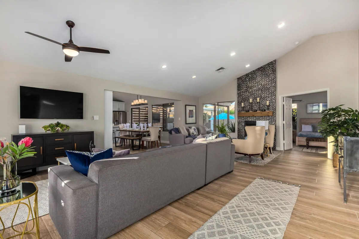 A spacious, open-concept living area featuring a large sectional sofa arranged around a modern coffee table, a wall-mounted oversized TV, a cozy fireplace, and an adjacent dining area. The space is filled with natural light and designed for relaxation and social gatherings.