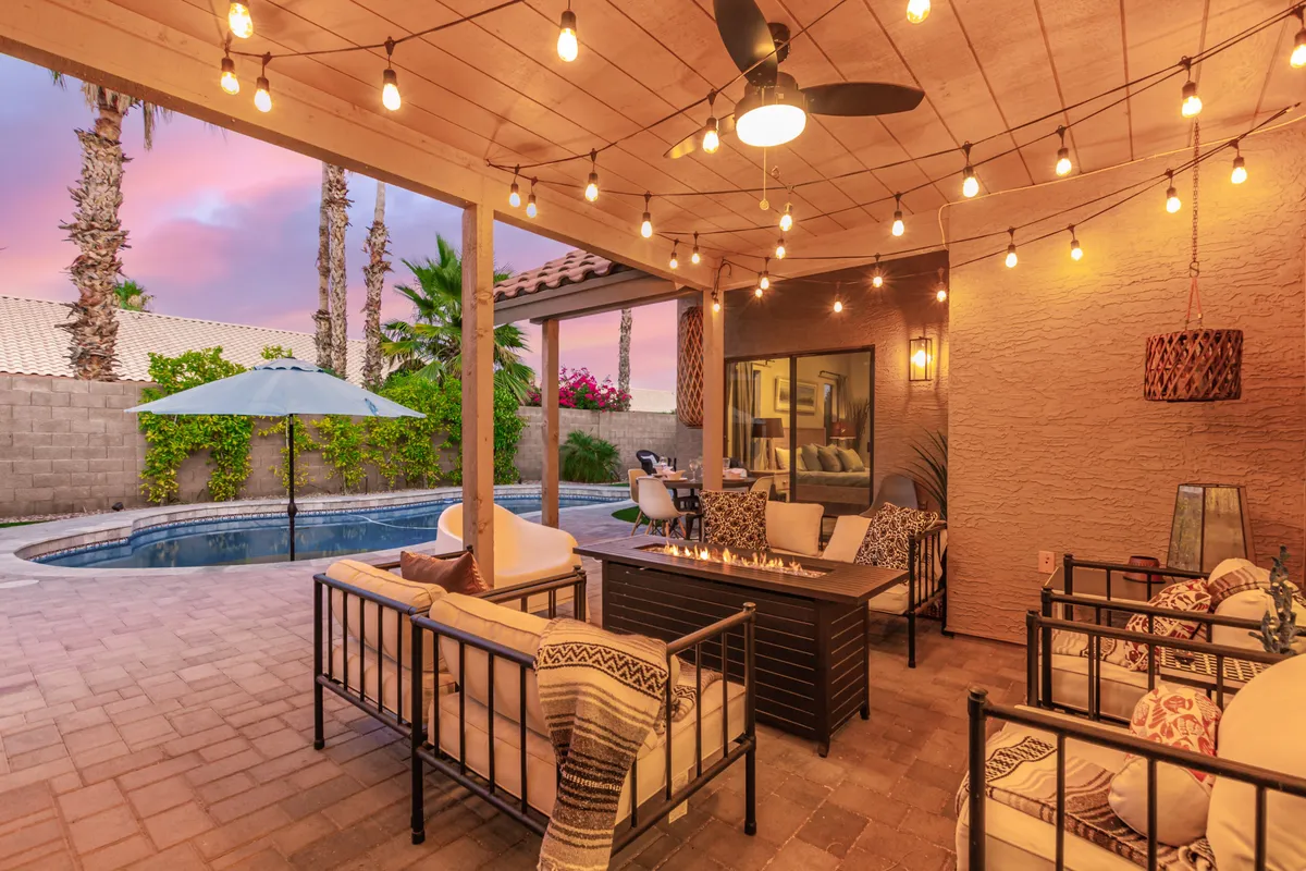 A cozy backyard setup with a glowing fire pit, comfortable outdoor seating, string lights overhead, and the edge of the swimming pool visible. Greenery frames the space, creating an inviting and magical environment for evening gatherings.