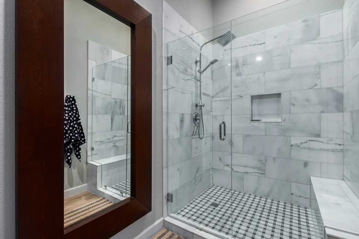 A stylish bathroom showcasing a glass walk-in shower, marble tiles, a chic mirror, and a patterned rug. The space exudes an elegant spa-like feel, underscoring comfort and luxury.